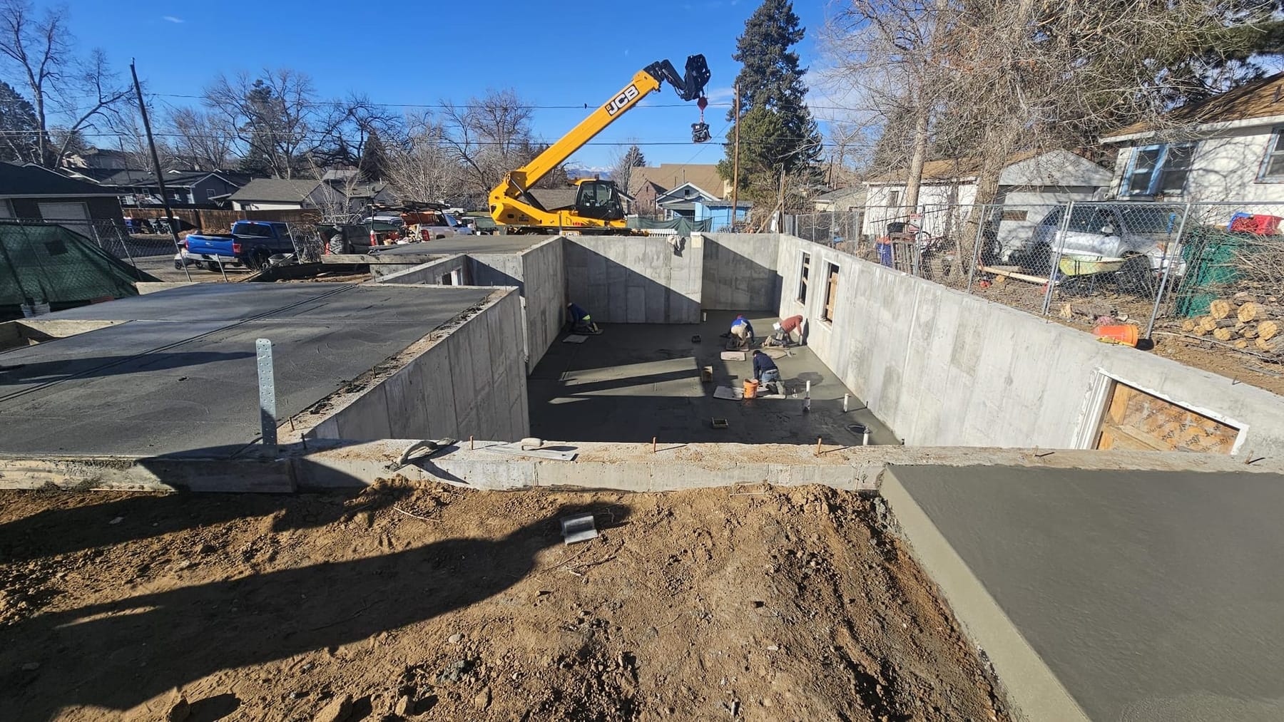 A dug and poured basement foundation on a Littleton lot, with excavated ground in the foreground and a telehandler working over the walls